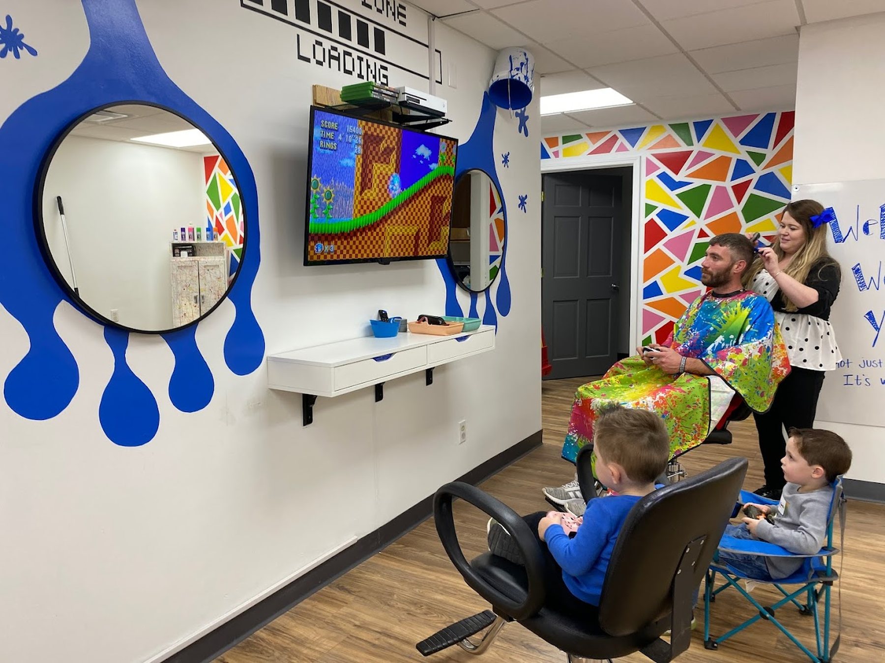 The salon interior: a wall painted with enormous blue paint drips with round mirrors set inside them, two boys on facing game stations, and a man in a rainbow splatter cape having his hair cut.