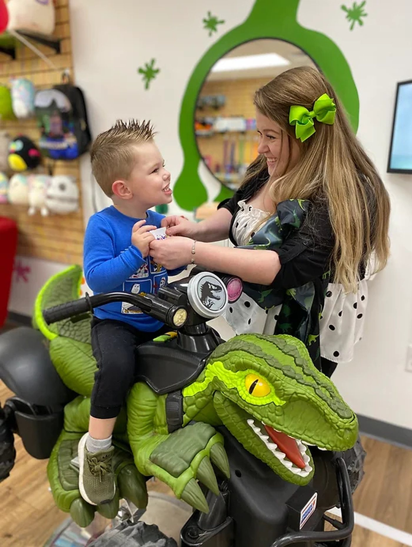 A boy sitting on a large green ride-on dinosaur, laughing with his stylist, against a wall painted with a green splat.